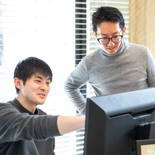 2人の男性社員がPCの画面を見ながら会話をしている様子